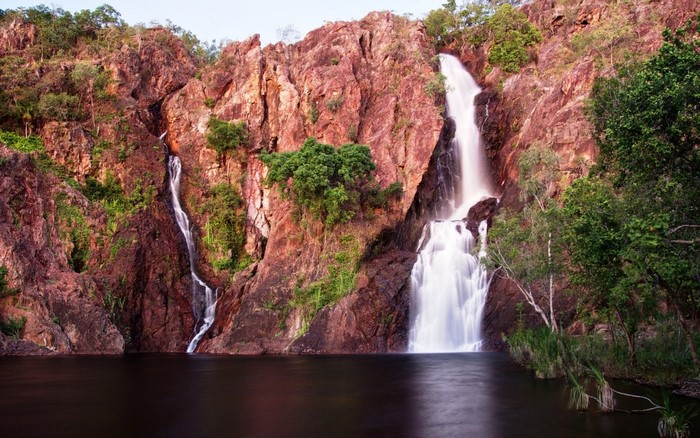 Twin waterfalls cascade down red rocky cliffs into a dark pool of water.