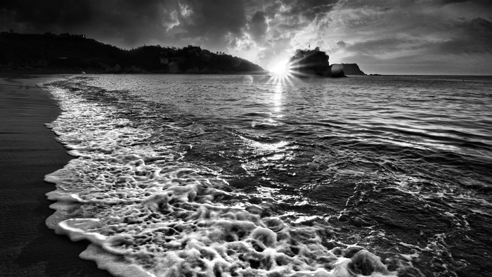 Monochrome sea with waves crashing on a sandy shore at sunset