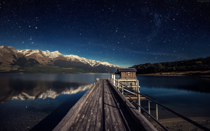 Wooden pier under starry sky with snow-capped mountains reflected in lake