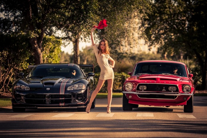 Woman posing between a black Dodge Viper and a red Ford Mustang