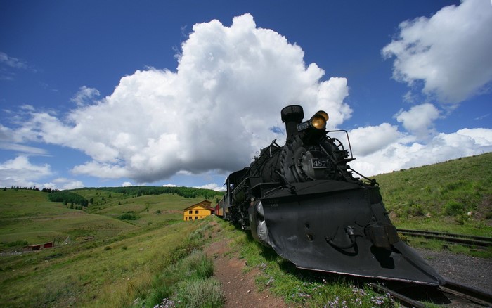 Steam locomotive on a grassy hill with a yellow building