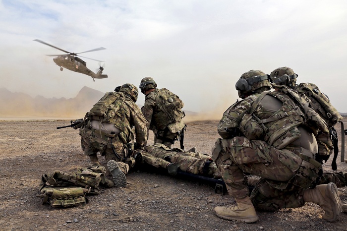 Soldiers preparing to load a casualty onto a stretcher as a helicopter approaches in a desert.