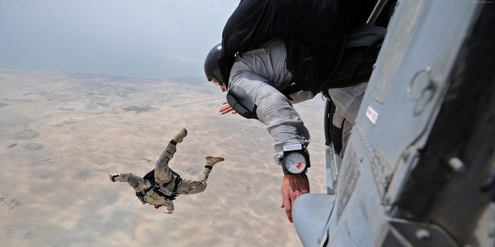 Two paratroopers jump from a military aircraft over a desert landscape.