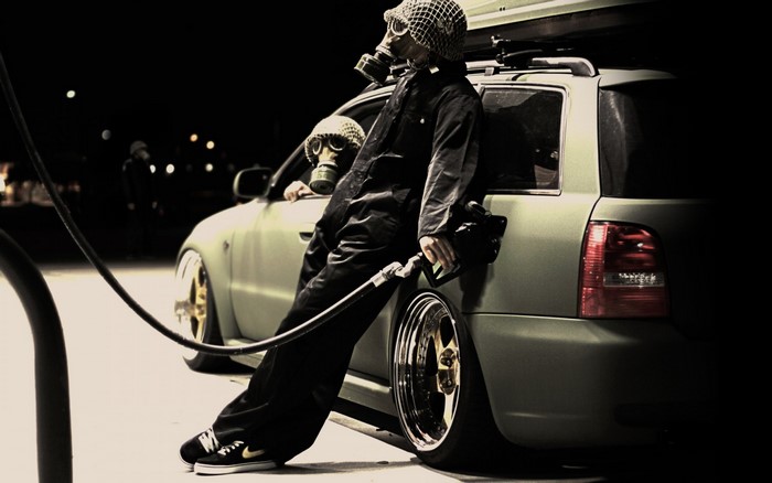 Two people in gas masks at a gas station with a car