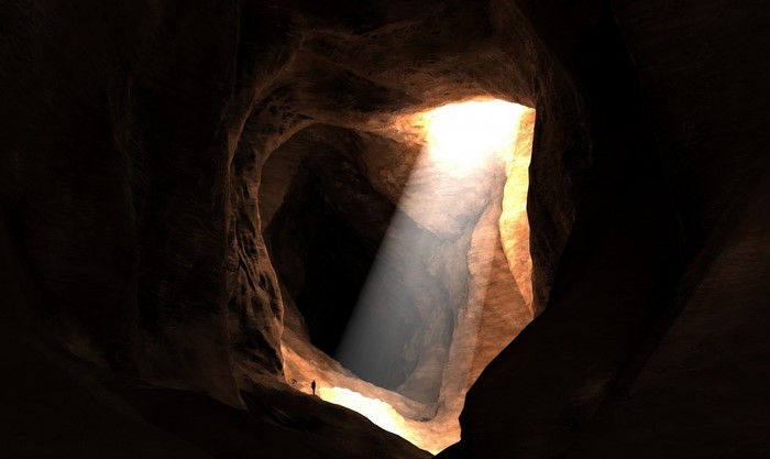 A lone figure stands in a dark cave with a sunbeam.