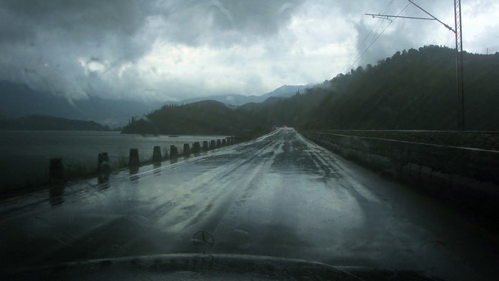 Wet road by a lake with mountains under stormy clouds