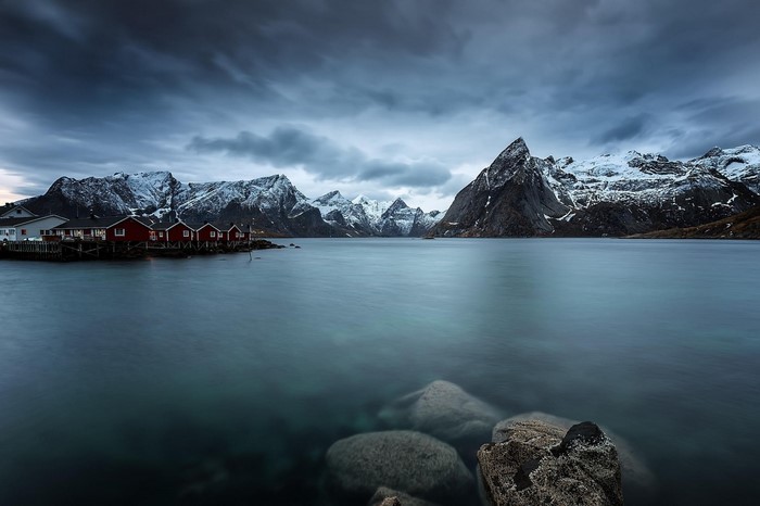 Red houses on a pier with snow-capped mountains and a cloudy sky