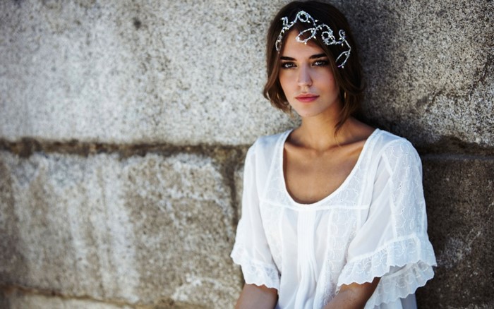 Woman with headpiece wearing white dress leaning against stone wall