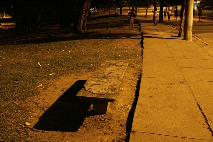 Concrete bench in a park at night with people walking on a sidewalk