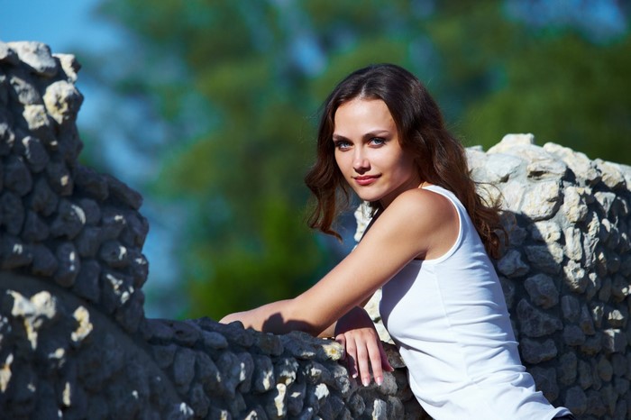Woman with long brown hair and blue eyes leaning on a stone wall