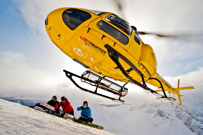 Yellow helicopter with Last Frontier Heliskiing logo over snowboarders