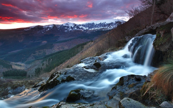 Waterfall cascading down rocks with snow-capped mountains and sunset sky