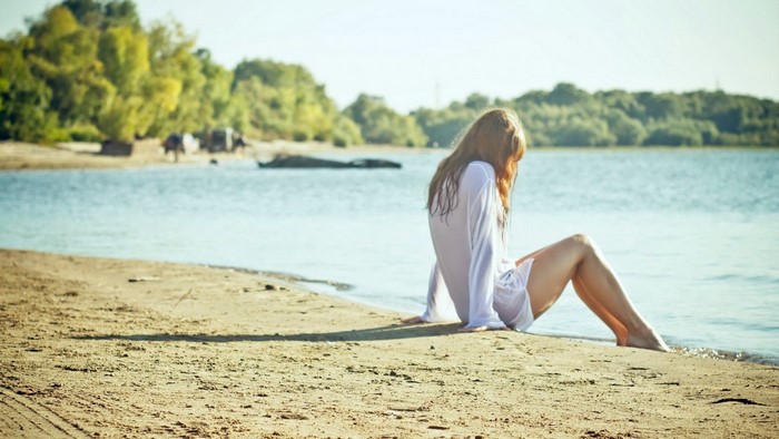 Woman in white shirt sitting on a sandy beach by the water