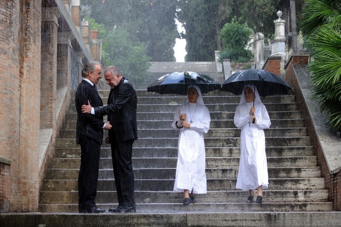 Two men in suits shake hands in the rain with nuns holding umbrellas