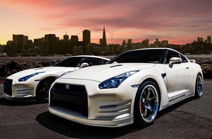 Two white Nissan GT-R sports cars with city skyline at sunset