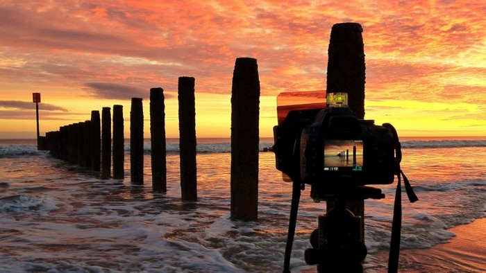 Camera photographing a sunset over the sea with wooden posts