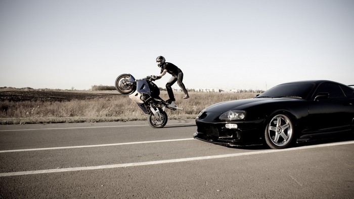 Motorcyclist doing a wheelie next to a black Supra