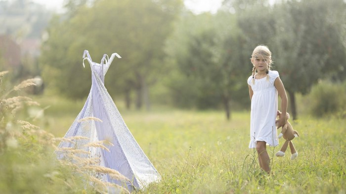 Girl in white dress with teddy bear near teepee in meadow