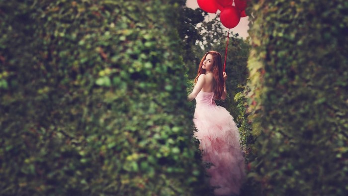Woman in pink dress holding red balloons in a hedge maze
