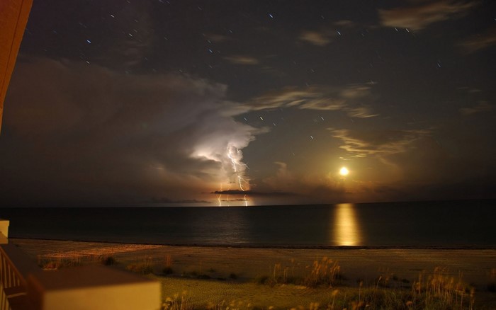 Lightning strikes over the ocean at night with a full moon.