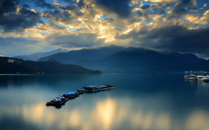 Boats docked on a calm lake with mountains and clouds