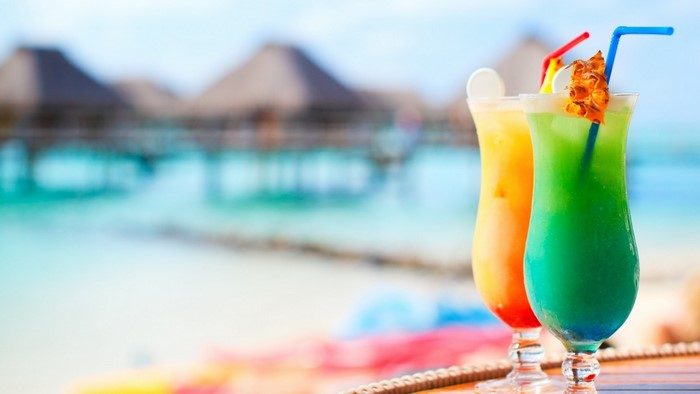 Two colorful cocktails on a table with a beach resort in the background