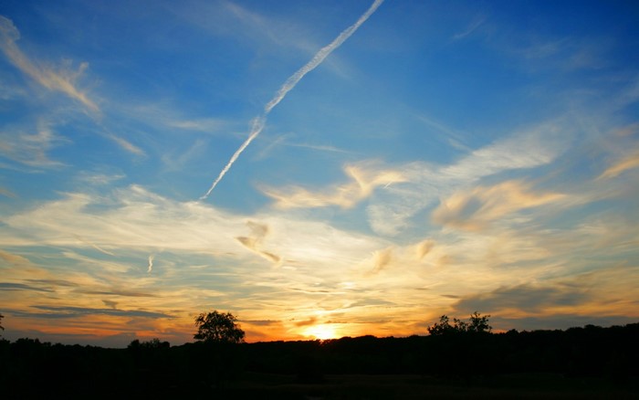 Sunset sky with clouds and contrail over silhouetted landscape