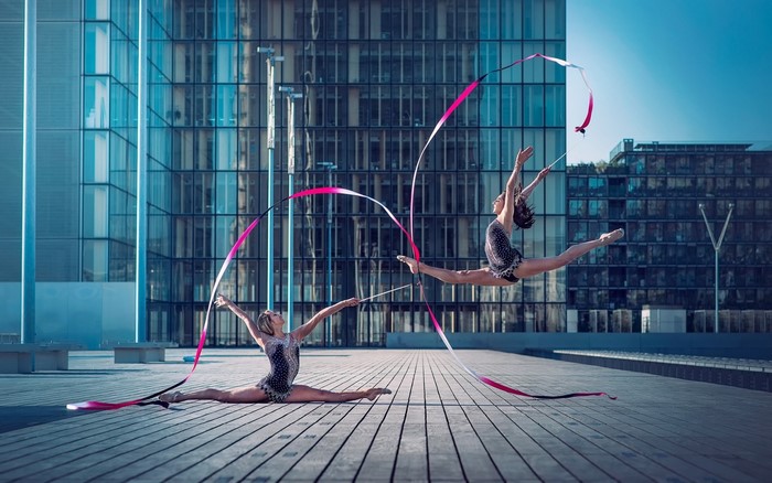 Two rhythmic gymnasts performing with ribbons in front of a modern building