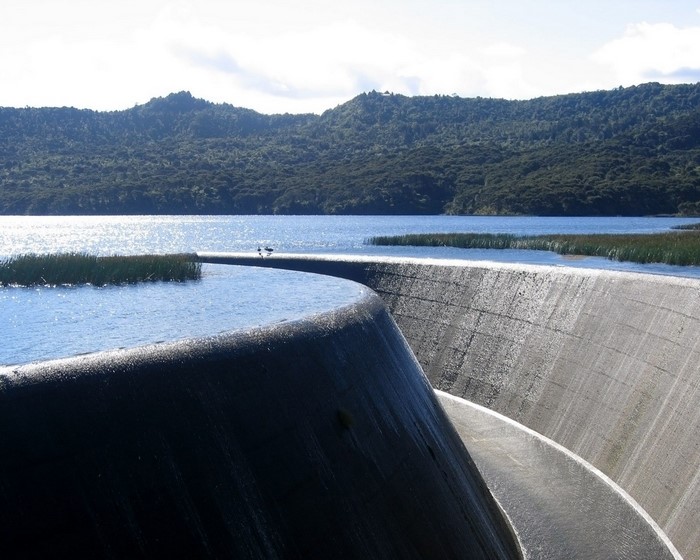 Curved concrete dam holding back a blue lake with forested hills