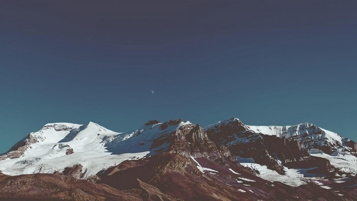 Snow-capped mountains under a clear blue sky with a crescent moon