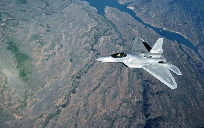 F-22 Raptor fighter jet flying over rugged terrain and water