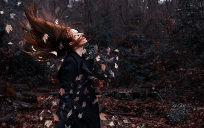 Redhead woman with long hair in a black coat in a forest with falling leaves