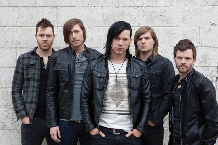 Five men in leather jackets and casual wear standing against a wall