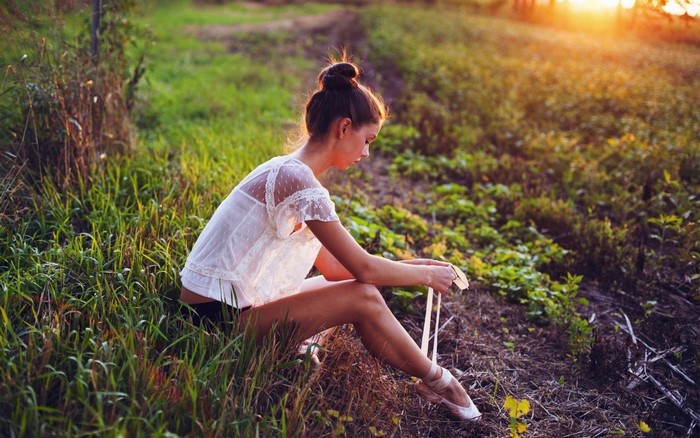 Woman in ballet attire tying pointe shoes in a field at sunset