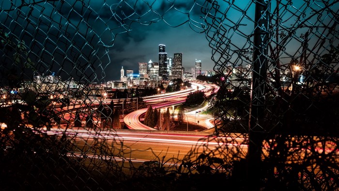 Night city skyline with light trails through a chain-link fence