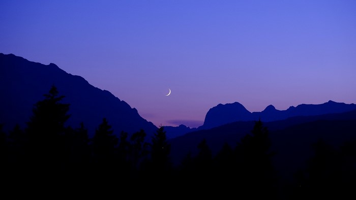 Crescent moon over silhouetted mountains and trees at dusk