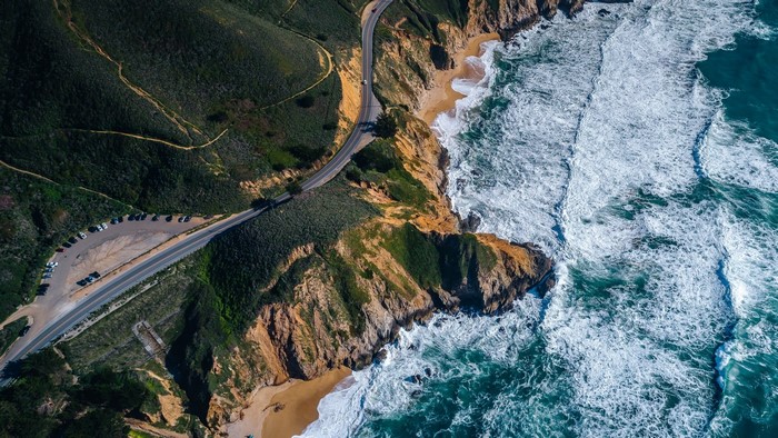 Aerial view of a winding road along a rugged coastline with crashing waves
