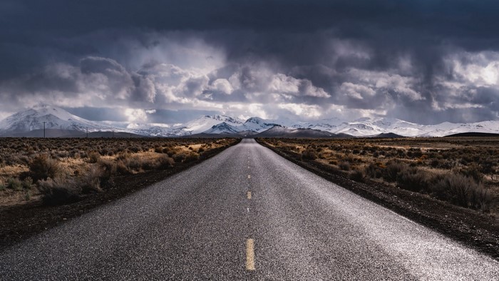 Empty road leading to snow-capped mountains under stormy clouds