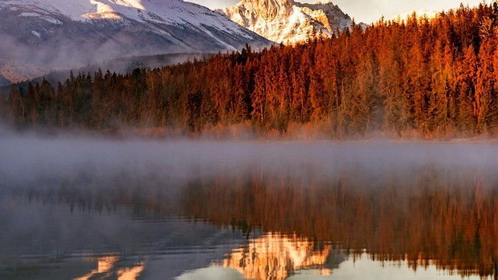 Misty lake reflecting snow-capped mountains and autumn forest