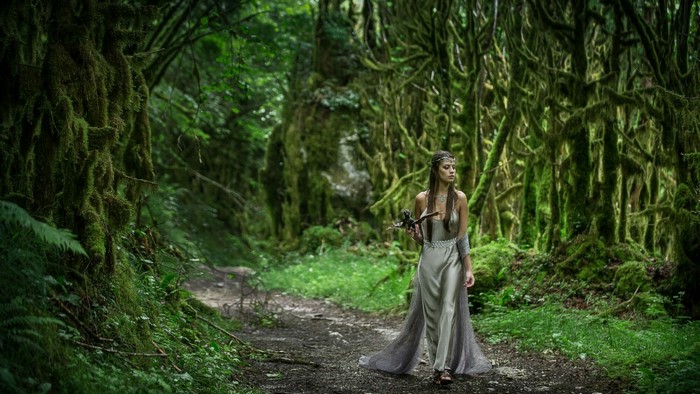 Woman in a silver dress holding a prop weapon in a mossy forest