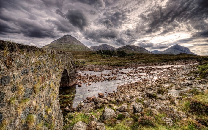 Stone bridge over a rocky river with mountains under cloudy sky