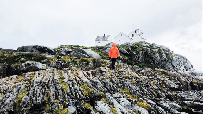 Person in orange jacket on rocky hill near white houses in Norway