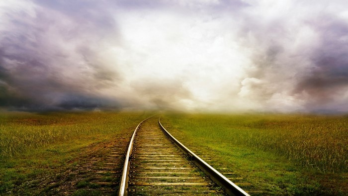 Railway tracks through a grassy field under a cloudy sky