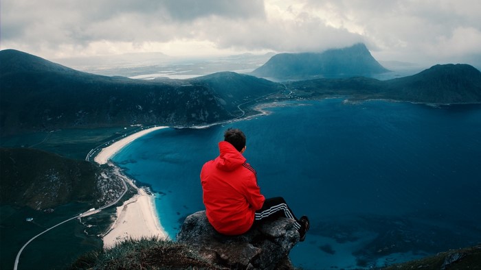 Man in red jacket sitting on cliff overlooking ocean and mountains