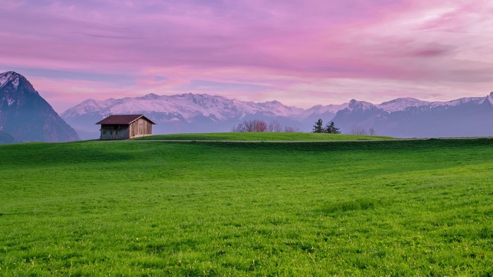 Wooden cabin on a grassy hill with snow-capped mountains and pink sky