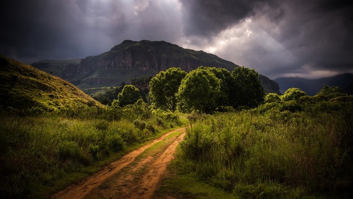 Dirt road through a grassy field with mountains under stormy clouds