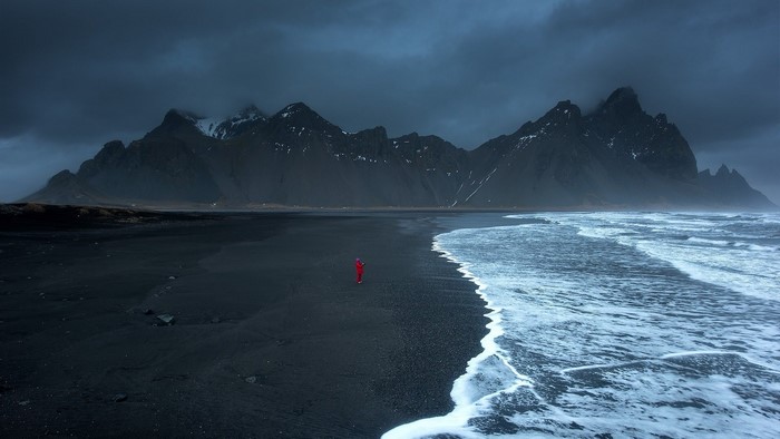 Person in red on black sand beach with Vestrahorn mountains, Iceland