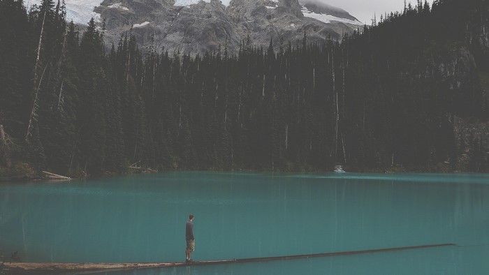 Man standing on a log in a turquoise lake with mountains and forest