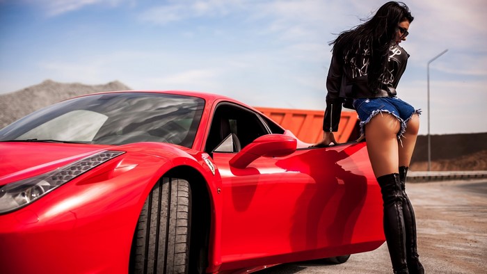 Woman in denim shorts and leather jacket leaning on a red Ferrari 458 Italia
