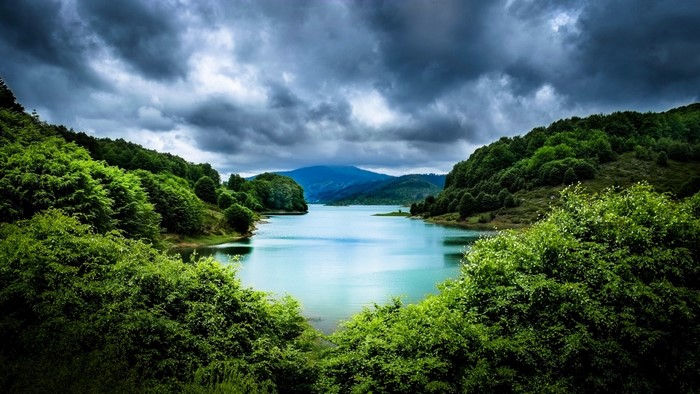 Lake surrounded by green trees and mountains under cloudy sky